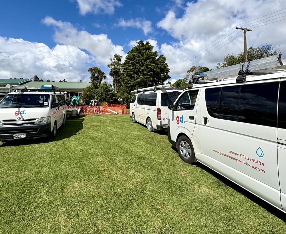 Four GD Plumbing Services vans lined up on the grass, the nearest carrying the company logo, phone number and website across its side.