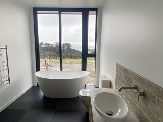 A freestanding bath set in front of a full height window looking out across paddocks to the hills, with dark large-format floor tiles and a heated towel rail beside it.