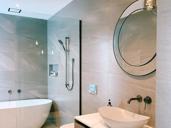 A bathroom with a freestanding bath beside a glazed walk-in shower, large stone-look wall tiles and a timber vanity with a white vessel basin.