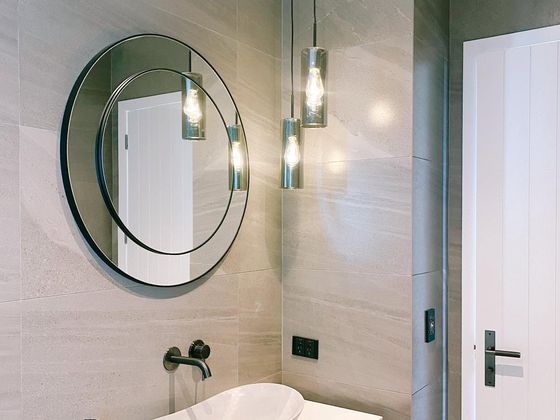A timber vanity with a white vessel basin and black wall-mounted tapware under a large round mirror, lit by three hanging glass pendants.