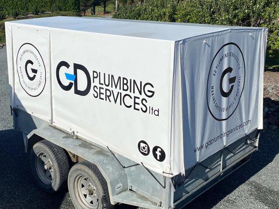 A GD Plumbing Services trailer with a white canvas cover carrying the company logo, parked on a gravel drive in front of a hedge.