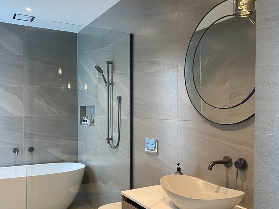 A grey tiled bathroom with a freestanding bath behind a glass screen, a shower on a rail, and a timber vanity with a stone vessel basin under a large round mirror.