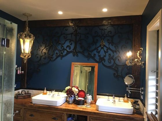 A deep navy bathroom with two white basins set on an antique timber vanity, ornate mirrors and a wrought iron wall panel behind.