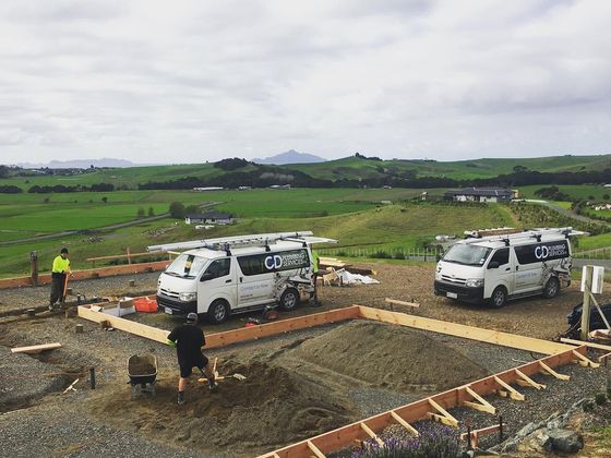 Two GD Plumbing Services vans at a new build, with timber floor framing laid out on the site and farmland running back to the hills.