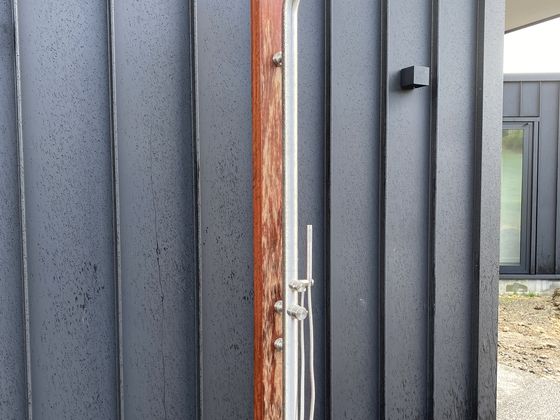 A stainless steel outdoor shower mounted on a hardwood backboard against the dark corrugated cladding of a new build.