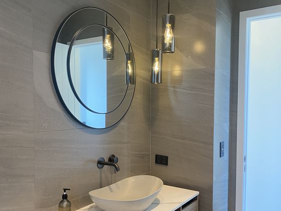 A white vessel basin on a timber vanity with black wall-mounted tapware, below a large round mirror and three hanging glass pendant lights.
