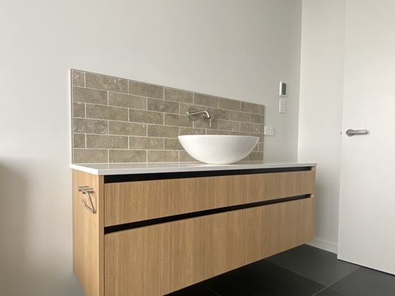 A wall-hung timber vanity with a white vessel basin and a chrome mixer against a brick-look tiled splashback, over dark large-format floor tiles.