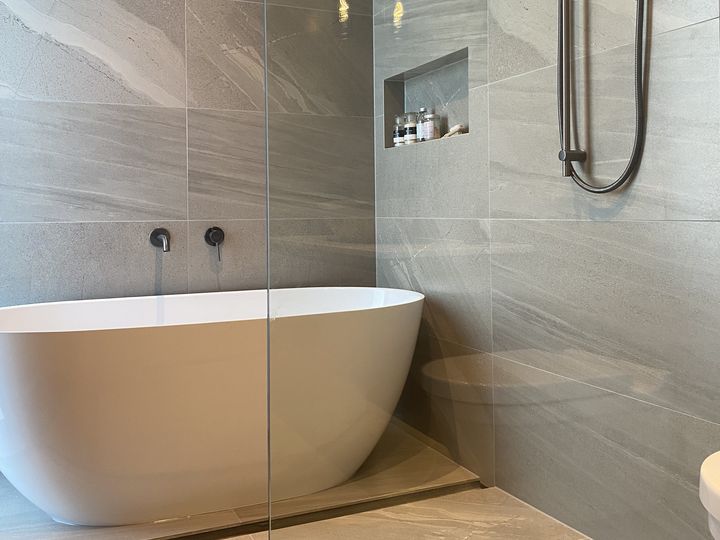 A freestanding bath beside a glazed walk-in shower in a bathroom tiled floor to ceiling, with black wall-mounted tapware, a hand shower on a rail and a tiled recess.