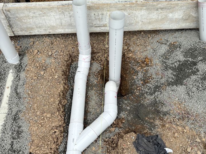 Two white PVC drainage risers and a swept junction set in the trench against the timber boxing of a new build foundation, before the slab is poured.