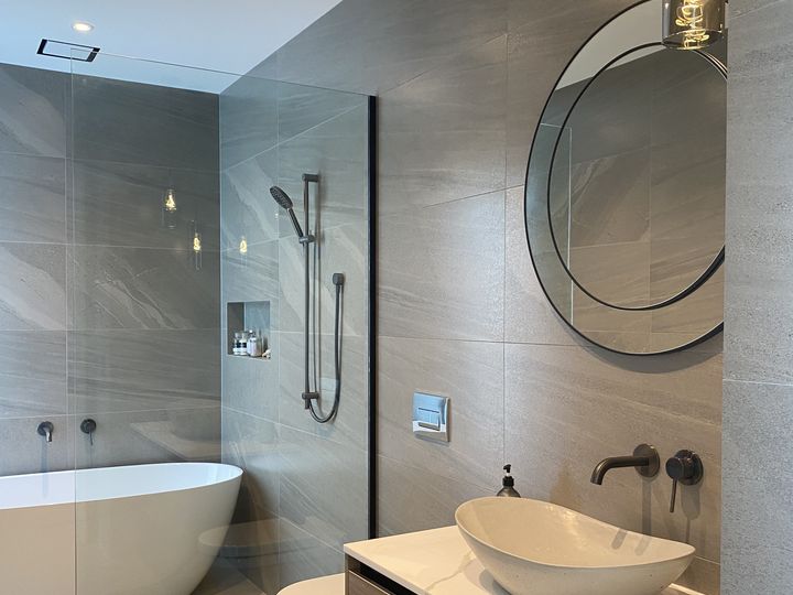 A finished bathroom with a freestanding bath behind a glass shower screen, a shower on a rail, and a timber vanity with a stone vessel basin under a large round mirror.